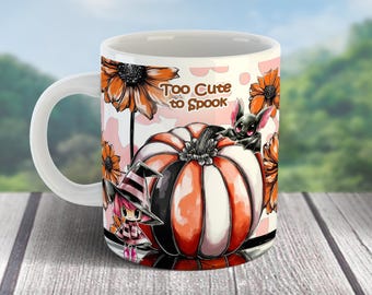 May include: White ceramic mug with a Halloween theme. The mug features a pumpkin, flowers, and a bat illustration. Text on the mug reads "Too Cute to Spook". The mug has a white handle and is set against a blurred background of a wooden surface and greenery.
