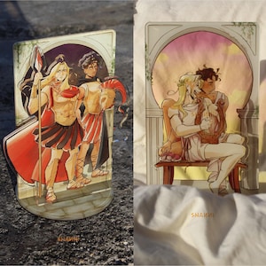 May include: Two acrylic standees featuring illustrations of classical figures. One depicts three figures in Roman attire, with a spear and helmet. The other shows two figures, one playing a lyre. The artist's signature, "SHANNI", is visible.