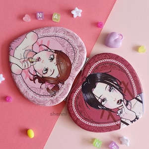 Könnte beinhalten: Zwei herzförmige Kompaktspiegel mit unterschiedlichen Designs. Ein Spiegel zeigt einen rosa Hintergrund mit einer Comic-Frau in einem rosa Kleid, die einen Lippenstift hält. Der andere Spiegel hat einen roten Hintergrund mit einer Comic-Frau in einem schwarzen Outfit, die sich einen Finger an die Lippen hält. Beide Spiegel haben einen weißen Spitzenrand.