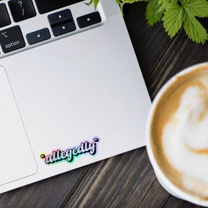 K&ouml;nnte beinhalten: Ein Laptop mit einem Regenbogen-Aufkleber, auf dem "allegedly" steht, auf der rechten oberen Ecke des Trackpads. Rechts im Bild ist eine Tasse Kaffee mit Latte Art.