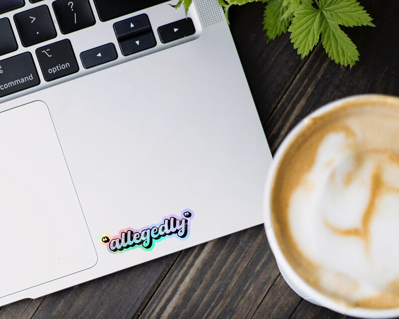 K&ouml;nnte beinhalten: Ein Laptop mit einem Regenbogen-Aufkleber, auf dem "allegedly" steht, auf der rechten oberen Ecke des Trackpads. Rechts im Bild ist eine Tasse Kaffee mit Latte Art.
