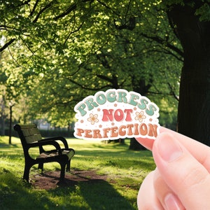 May include: A sticker with a retro design featuring the text "Progress Not Perfection" in a colorful font with flowers. The sticker is being held in a hand against a backdrop of a green park with a bench.