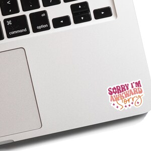 May include: A pink and white sticker with the text "Sorry I'm Awkward Sorry" on a silver laptop.