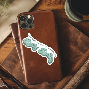 May include: A brown leather phone case with a sticker that says "You guys go ahead, I'm gonna stay" in a blue and green retro font.
