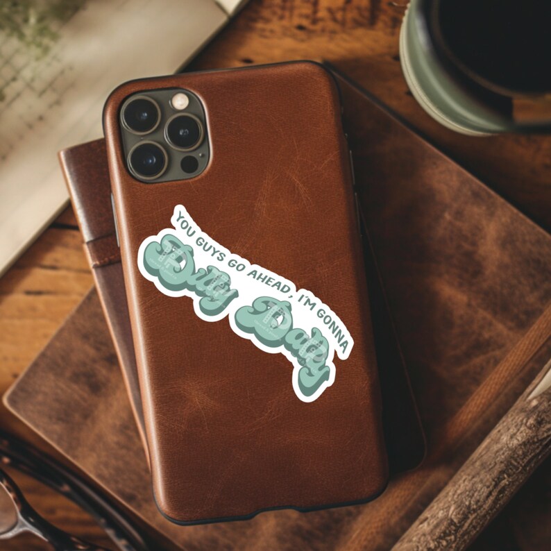 May include: A brown leather phone case with a sticker that says "You guys go ahead, I'm gonna stay" in a blue and green retro font.