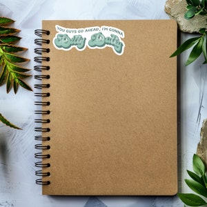 May include: A brown notebook with a spiral binding and a green sticker that says "You guys go ahead, I'm gonna dilly dally".
