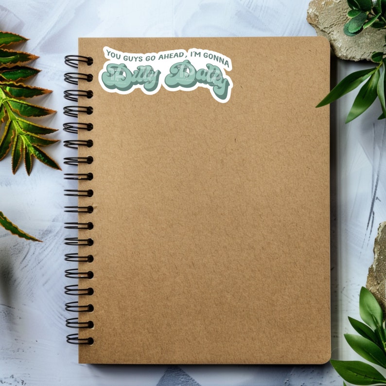 May include: A brown notebook with a spiral binding and a green sticker that says "You guys go ahead, I'm gonna dilly dally".