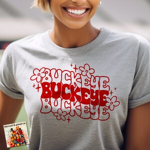 May include: Gray t-shirt with a red and white graphic design that says "Buckeye" three times. The graphic is surrounded by white flowers and plus signs.