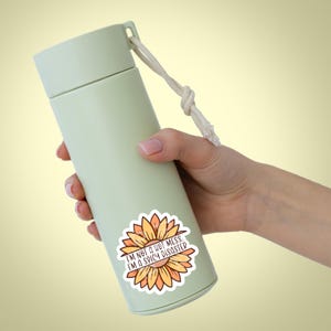 May include: A green reusable water bottle with a white rope handle and a sticker that says "I'm not a hot mess. I'm a spicy disaster." The sticker features a yellow and orange sunflower.