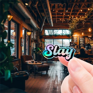 May include: A holographic sticker with the word "Slay" in a bold, cursive font. The sticker is iridescent and has a rainbow effect. The sticker is being held by a hand in a dimly lit cafe setting.