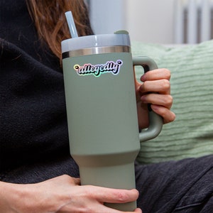 K&ouml;nnte beinhalten: Eine gr&uuml;ne Reise-Tasse mit Henkel und Strohhalm. Die Tasse hat einen Aufkleber mit der Aufschrift "angeblich" in Regenbogenfarben.