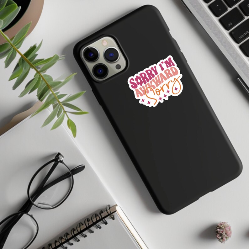May include: Black phone case with a pink, orange, and red sticker that says "Sorry I'm Awkward Sorry" with stars.