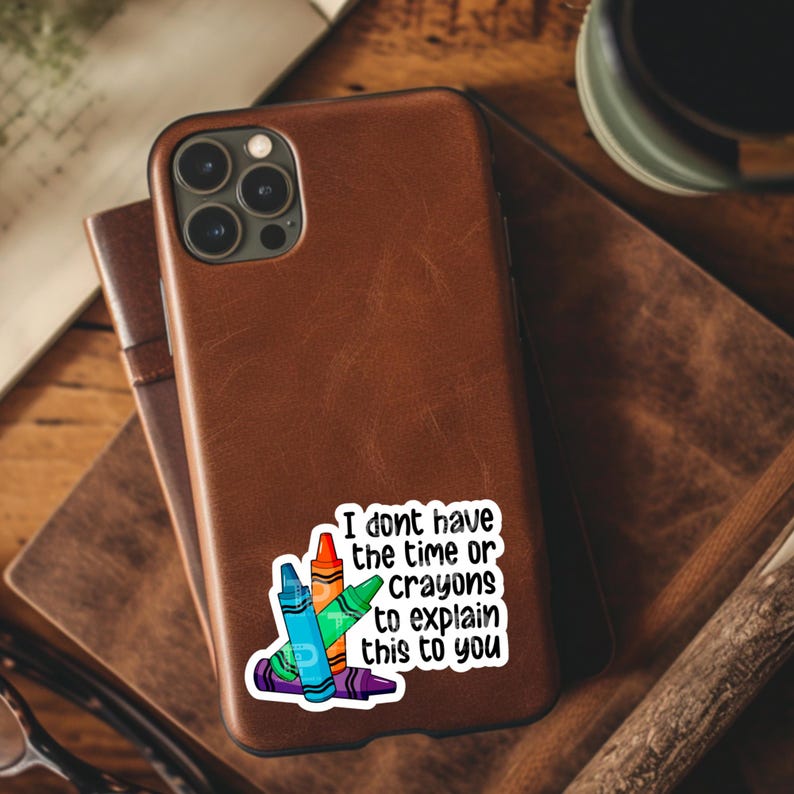 May include: A brown leather phone case with a sticker that says "I don't have the time or crayons to explain this to you". The sticker features a stack of colourful crayons.