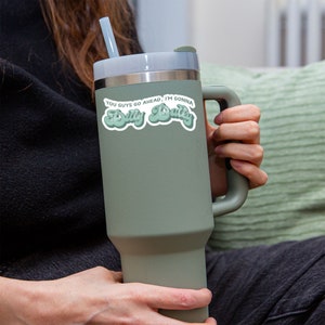 May include: A green travel mug with a white sticker that says "You guys go ahead, I'm gonna "Dilly Dally" ".