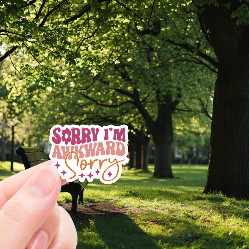 May include: A pink and white sticker with the text "Sorry I'm Awkward Sorry" in a retro font. The sticker is held in a hand in front of a green grassy area with trees in the background.