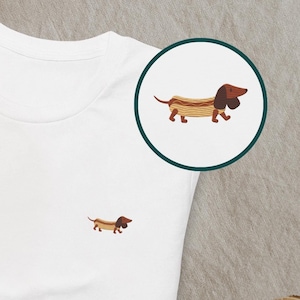 May include: White t-shirt with a small embroidered dachshund design on the front. The dachshund is brown and tan, with a hot dog bun design. A larger version of the design is in a circle on the top right.