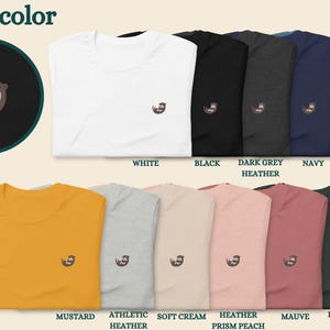 May include: A variety of t-shirts in different colors, featuring a small embroidered otter design. The colors include white, black, dark grey heather, navy, heather deep teal, mustard, athletic heather, soft cream, heather prism peach, mauve, and heather forest.