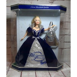 May include: Special Millennium Edition Barbie doll in a display case. The doll wears a dark blue gown with silver accents and a tiara. The case has a blue and silver trim, with the text "Millennium Princess" and "Barbie". A Millennium Keepsake is included.