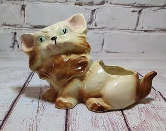 May include: A vintage ceramic cat planter in cream, brown, and tan. The cat is in a seated position with a long tail and a planter opening on its back. The cat has green eyes and a detailed fur design.