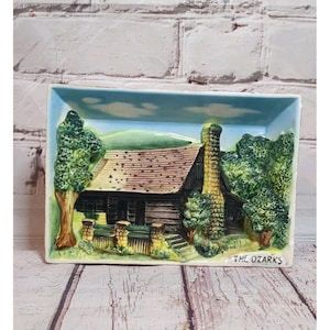 Könnte beinhalten: Eine rechteckige Keramikschale mit einem erhabenen Relief einer Hütte in den Ozark Mountains. Die Hütte hat ein braunes Dach und einen gelben Schornstein. Der Text "THE OZARKS" befindet sich unten.