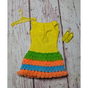 以下が含まれることがあります： 緑、オレンジ、青色の鉤針編みスカートが付いた黄色の人形用ドレス。 ドレスは黄色のハンガーと黄色の人形の靴のペアで表示されています。 ドレスは布と糸でできています。