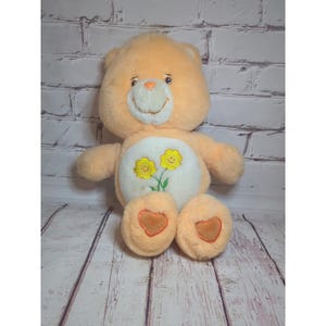 Könnte beinhalten: Ein pfirsichfarbener Care Bear-Plüschbär mit einem lächelnden Gesicht und einem weißen Bauch mit zwei gelben Blumen. Der Bär hat herzförmige Akzente auf seinen Pfoten. Der Hintergrund ist eine weiße Backsteinmauer und eine Holzoberfläche.
