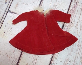 May include: A red velvet doll coat with short sleeves and a white faux fur collar. The coat is open in the front and has a flared skirt. The garment is likely vintage and is displayed on a light wood surface.