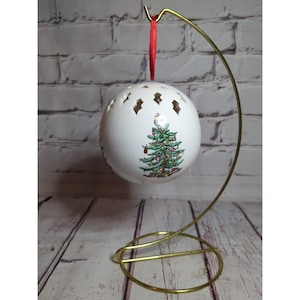 May include: A white Christmas ornament with a festive Christmas tree design and diamond-shaped cutouts. The ornament hangs from a red ribbon on a gold-colored metal stand. The tree is decorated with red and green accents.