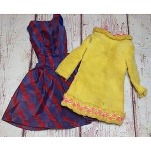 May include: Two vintage doll dresses. One is a blue and red striped dress with a pleated skirt. The other is a yellow coat with a decorative collar and pink and gold trim along the hemline. The dresses are on a light wood grain surface.