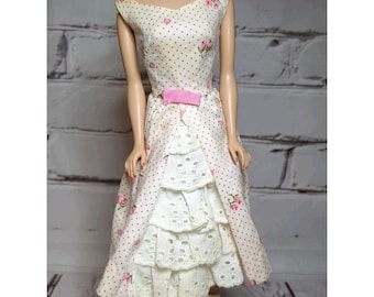 May include: A doll wearing a sleeveless dress with a cream background, small red polka dots, and pink floral accents. The dress features a pink bow at the waist and a layered white eyelet lace skirt with scalloped edges that extends below the hem.
