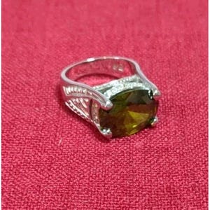 Op de afbeelding: Een zilveren ring met een grote, ovaal geslepen, olijfgroene edelsteen. De ring heeft een decoratief filigraanontwerp aan de zijkanten en een band van kleinere, heldere stenen. De ring is geplaatst tegen een rode achtergrond.