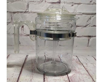Op de afbeelding: Een koffiezetapparaat van helder glas met een cilindrisch lichaam, een bijpassend glazen deksel met handvat en een stevig glazen handvat aan de zijkant. Een gepolijste metalen band omsluit het bovenste deel van de glazen kan.