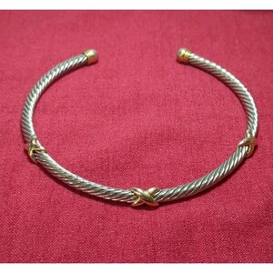 May include: Silver-tone and gold-tone twisted cable bracelet. The bracelet features gold accents at the ends and at the center, with a crisscross design. The jewelry is displayed on a red background.