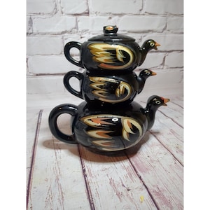 Może przedstawiać: Ułożony w stos czarny zestaw do herbaty z trzema częściami, z których każda ma kształt kaczki. Czajniki mają złote i pomarańczowe wzory skrzydeł i czerwone akcenty na dziobach. Zestaw stoi na drewnianej powierzchni.