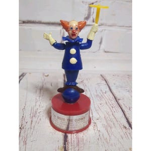May include: A vintage Bozo the Clown push button puppet toy. The clown figure has orange hair, a red nose, and is wearing a blue outfit with white gloves. The base is red with text that reads "Bozo the Clown".