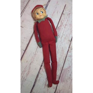 Op de afbeelding: Een pluche elfpop met een rood stoffen lichaam, lange armen en lange benen. De pop heeft een geschilderd gezicht met blauwe ogen, roze wangen en een glimlachende mond. Het draagt een bijpassende rode puntmuts met een groene kraagaccent.