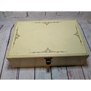 May include: A rectangular, cream-colored jewelry box with a decorative black floral design on the lid. The box has a metal clasp and is set against a weathered wooden background.