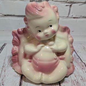 May include: A vintage ceramic figurine of a baby in a pink and cream color scheme. The baby is seated and holding a pink object, with rosy cheeks and a sweet expression. The figurine is surrounded by a ruffled, pink-edged blanket.