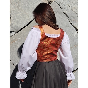 Pu&ograve; includere: Un gilet di broccato rosso e oro con scollo quadrato e bordatura di pizzo bianco. Il gilet &egrave; indossato sopra una blusa bianca a maniche lunghe con volant ai polsini.