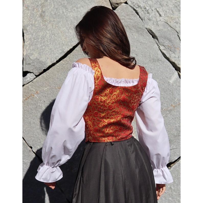 Pu&ograve; includere: Un gilet di broccato rosso e oro con scollo quadrato e bordatura di pizzo bianco. Il gilet &egrave; indossato sopra una blusa bianca a maniche lunghe con volant ai polsini.