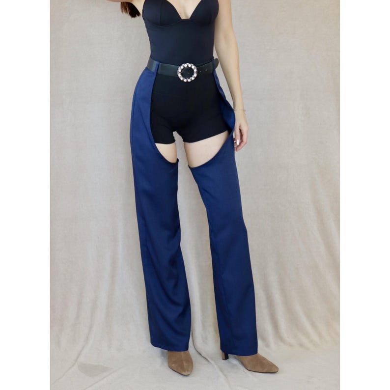 May include: A navy blue chaps-style pant with a high waist and open sides. The pants are worn with a black bodysuit and a black belt with a silver buckle. The model is wearing brown pointed-toe heels.