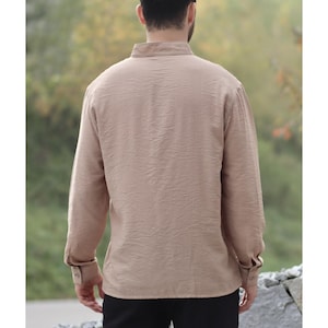May include: A tan long-sleeve shirt with a textured fabric. The shirt has a mandarin collar and buttoned cuffs. The back view of the shirt is shown, with the person wearing the shirt facing away from the camera. The shirt is made of a lightweight material.