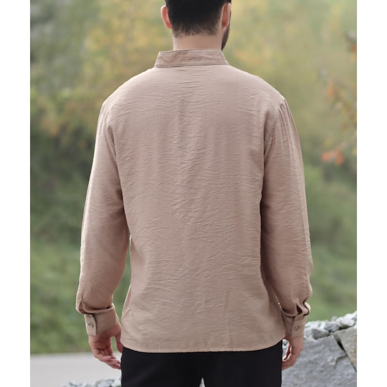 May include: A tan long-sleeve shirt with a textured fabric. The shirt has a mandarin collar and buttoned cuffs. The back view of the shirt is shown, with the person wearing the shirt facing away from the camera. The shirt is made of a lightweight material.