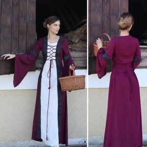 May include: A long, burgundy dress with a white under layer and black lace trim. The dress has a lace-up bodice and long sleeves. The model is holding a wicker basket.