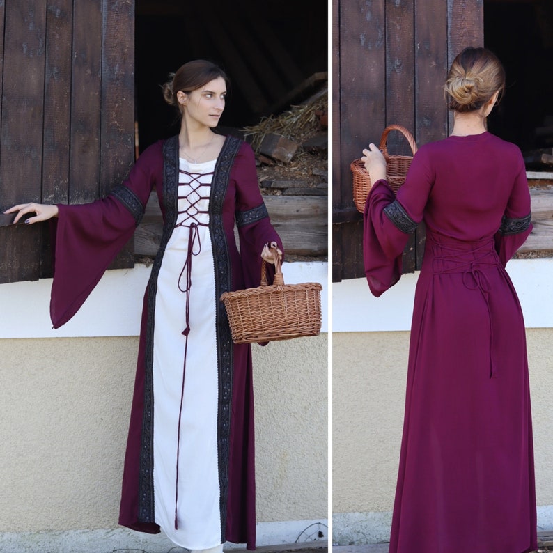 May include: A long, burgundy dress with a white under layer and black lace trim. The dress has a lace-up bodice and long sleeves. The model is holding a wicker basket.