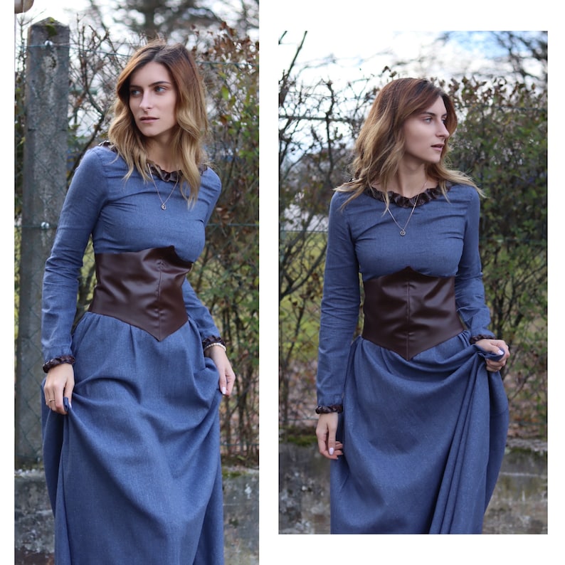 May include: A woman wearing a blue dress with a brown leather belt. The dress has long sleeves and a round neckline. The woman is standing in front of a green bush.