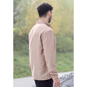 May include: A tan-colored long-sleeve shirt with a textured fabric. The shirt has a button-down collar and cuffs. The person in the image is wearing a black pair of pants. The background is blurred with green and brown tones.