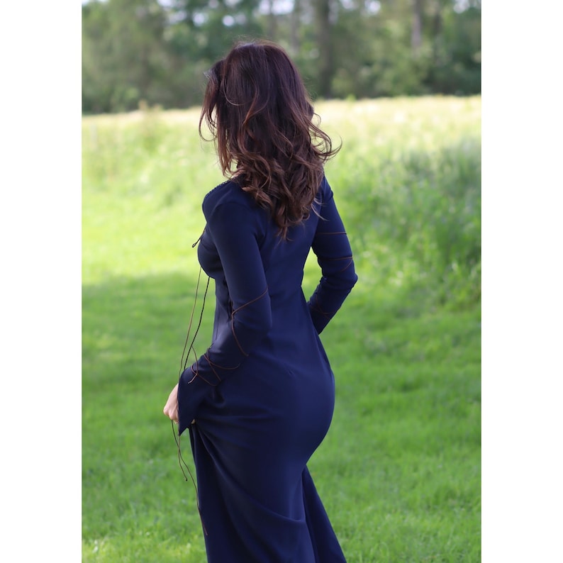 May include: A woman wearing a long, dark blue dress with brown trim. The dress has a lace-up front and long sleeves. She is standing in a field of green grass.