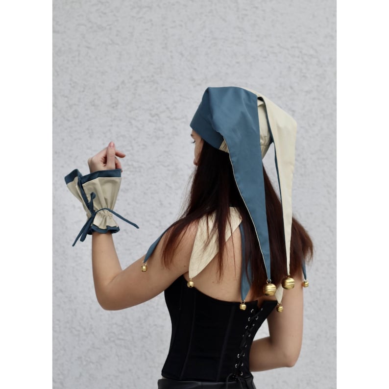 May include: A blue and cream-colored jester hat with long, pointed tips and small gold bells. Matching wrist cuffs with blue ties and cream-colored trim are also visible. The hat and cuffs are part of a costume or theatrical ensemble.