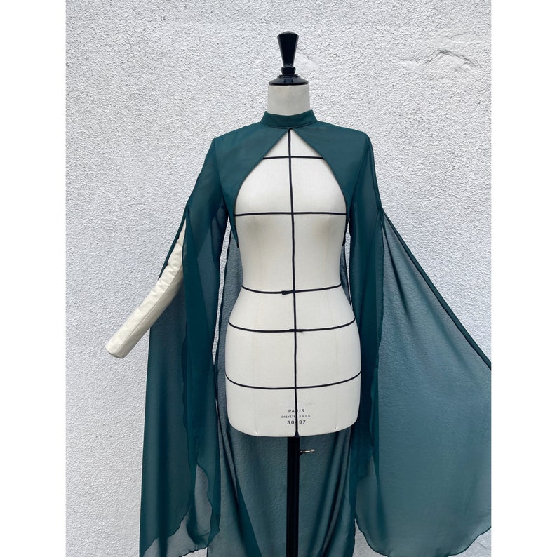 May include: A long, dark green sheer cape with a high neckline and long sleeves. The cape is open at the front and has a flowing, dramatic silhouette.